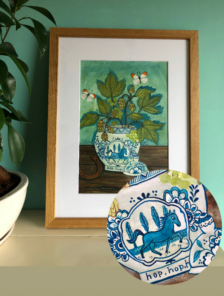 wilde bloemen stilleven schilderij in natuurlijke tinten als blauw, bruin en groen. Dit boeket staat in een Delfts Blauwe Vaas met een paard in galop. Op de tafel naast de blauw witte vaas ligt een hoefijzer. Dit bloem stilleven met hopplanten heeft een hoefijzer en twee vlindertjes. De vlinders zijn oranje tipjes.