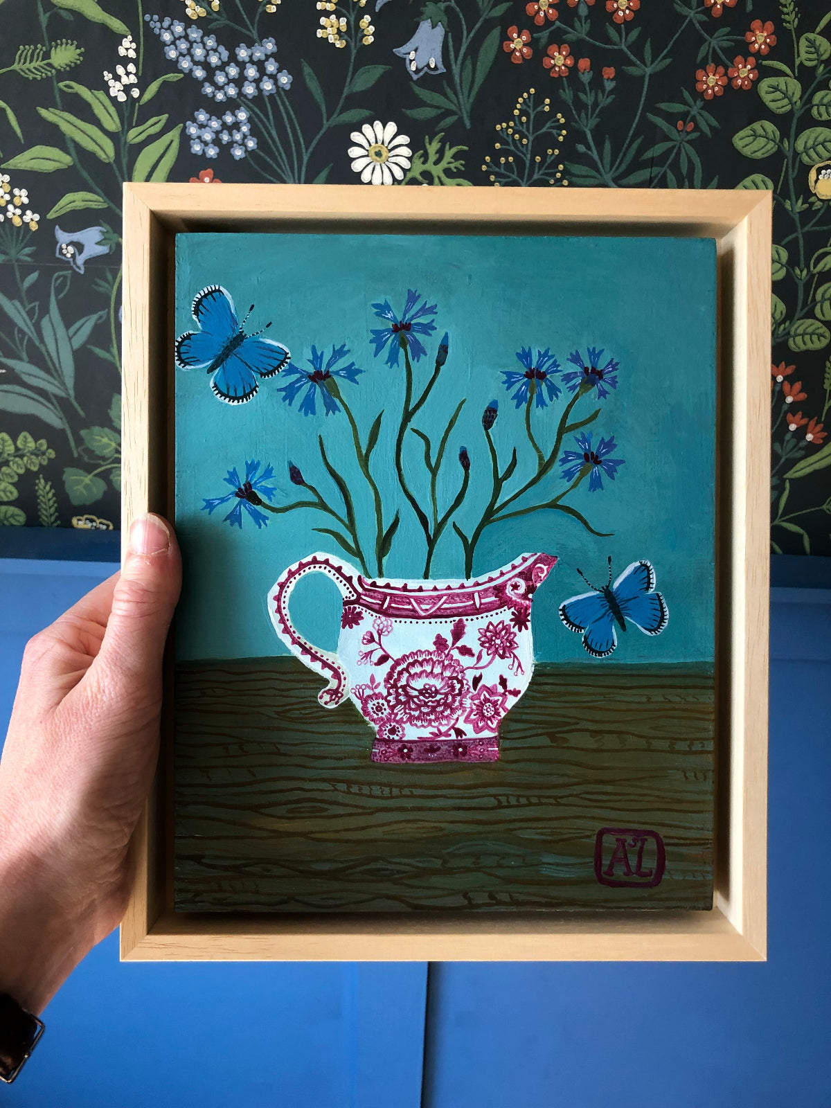 kleurrijk handgeschilderd korenbloem stilleven in blauwe tinten. De korenbloempjes staat in een burgenland  theekopje van villeroy en boch. Op dit handgemaakte schilderij staan ook twee blauwe vlindertjes. Dat zijn icarus blauwtjes.